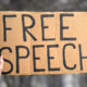 Hands hold up a cardboard "Free Speech" sign at protest rally