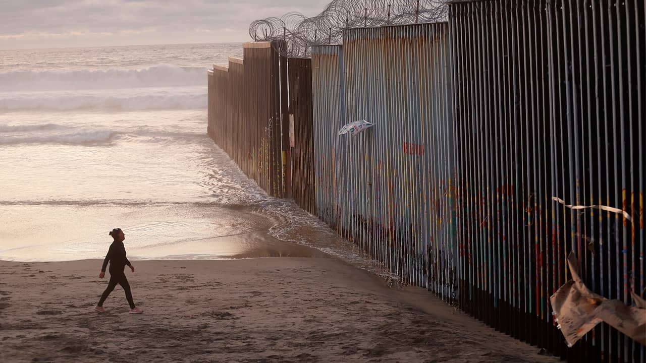 Photo of a woman walking along the beach next to the border wall in Mexico